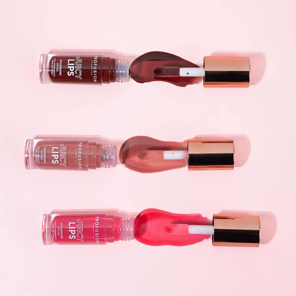 Three Profusion Cosmetics Juicy Lips lip tints in shades of deep red, mauve, and bright pink with rose gold caps are displayed on a pink background.