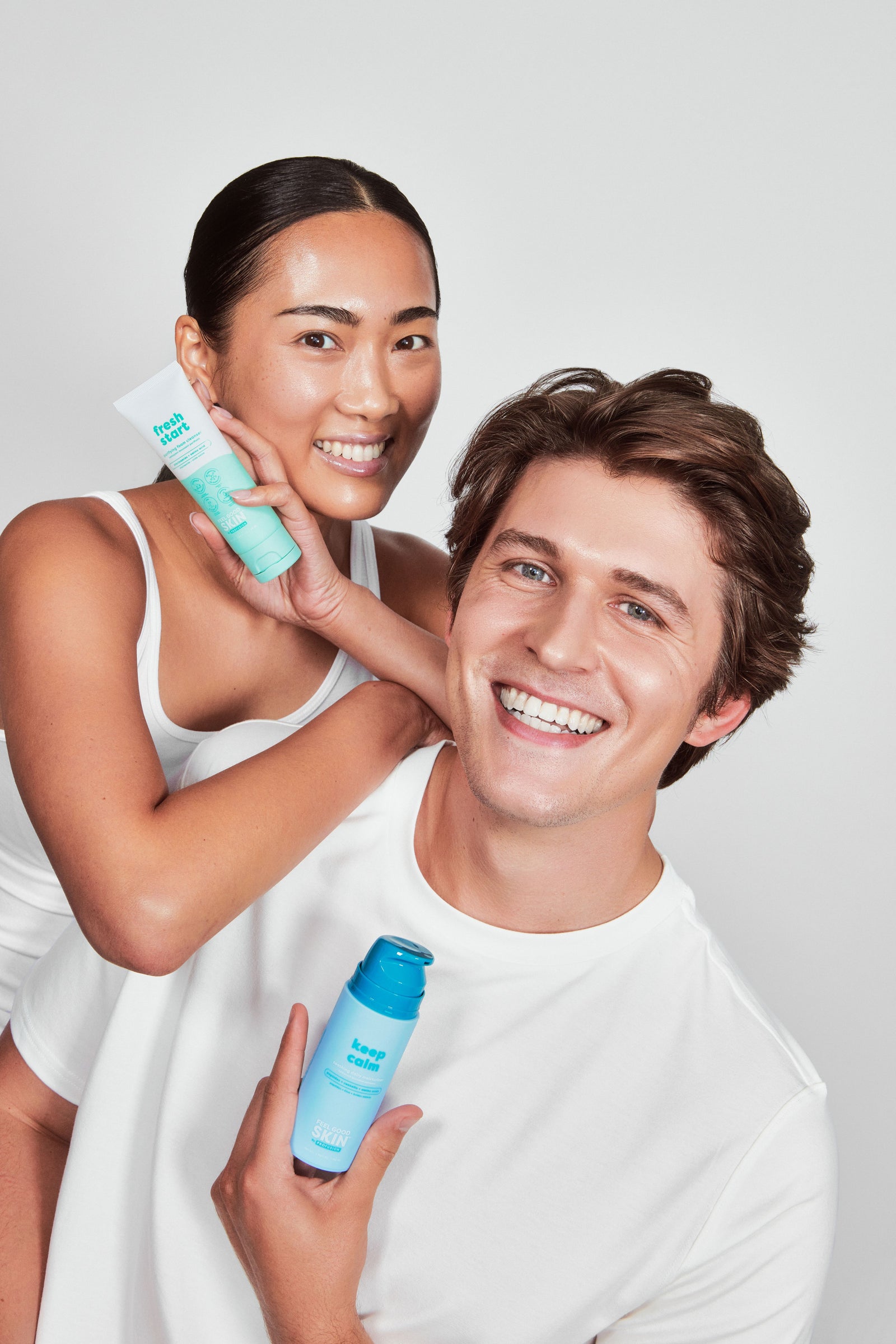 A smiling woman in a white tank top holds a light blue and white tube of hair product while standing behind a smiling man in a white t-shirt who holds a matching light blue bottle. They are posed against a solid light gray background.