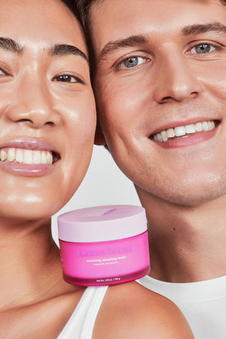 Two smiling people in a close-up beauty shot holding a bright pink jar labeled 'LUMIÈRE' in front of their faces against a light neutral background.