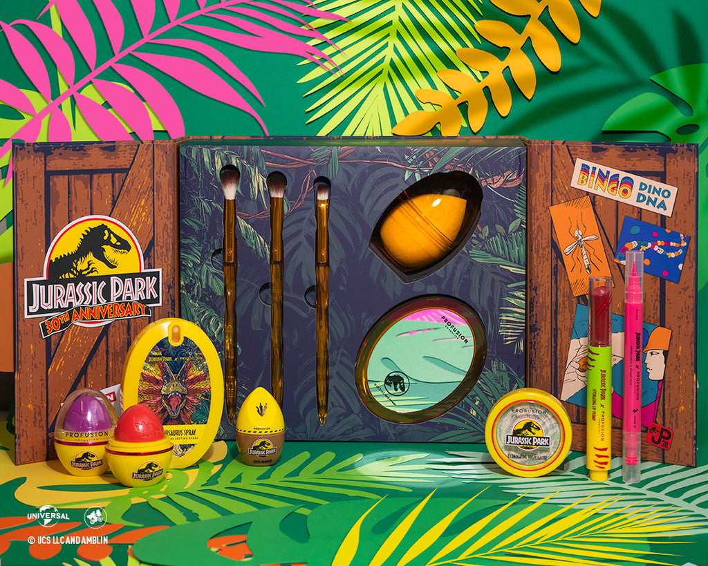 opened brush set with jurassic logo on left and three eyeshadow brushes and beauty blender and mirror, and on the right side different cartoon pictures one of a mosquito, a miner finding a mosquito in amber the words '' Bingo, Dino, DNA" Background with tropical leaves and for decoration in front of brush set there is the jurassic line lip balm, setting spray, bronzer, highlighter, lip plump and liner