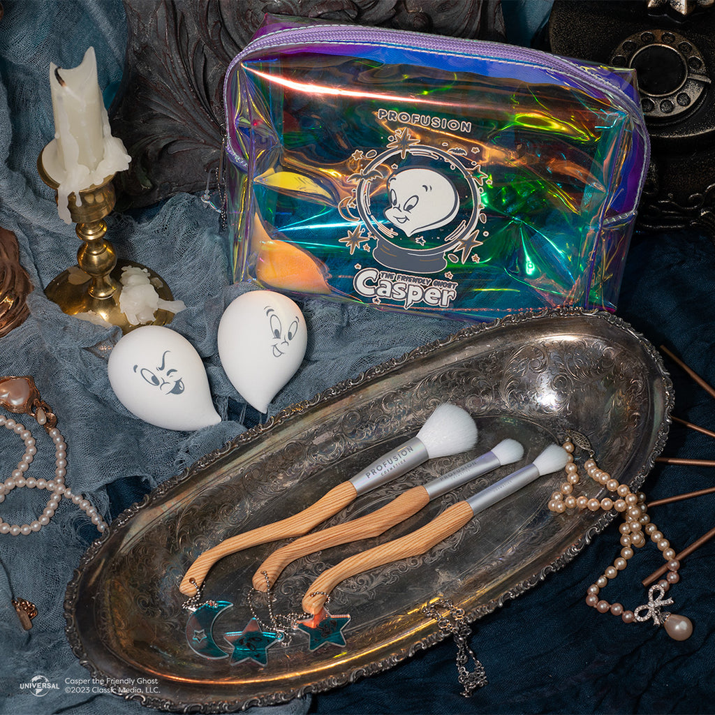 the iridescent bag with two casper face sponges and three makeup brushes laid out on a siler tray with a blue backdrop and pearl necklaces and white candle for decorations