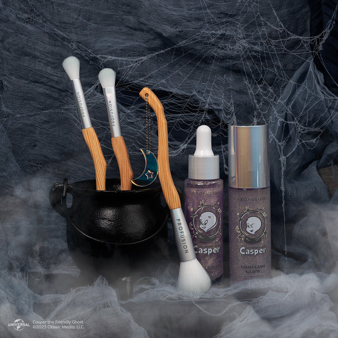 two makeup brushes in a small cauldron for decoration, and one more brush leaning against the cauldron and the face primer and setting spray to the right side of the brushes.