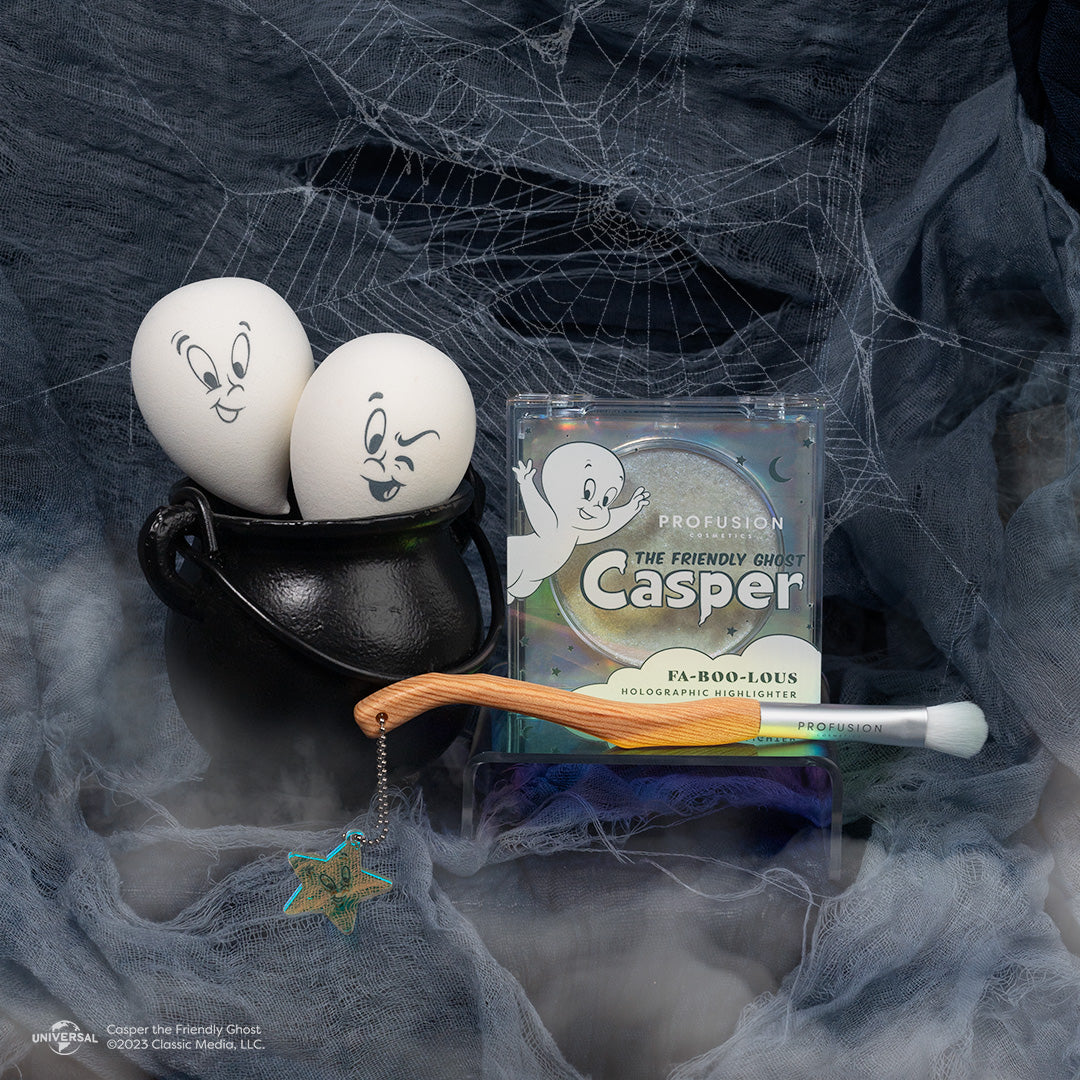 dark backdrop with spiderwebs and two casper face sponges in a cauldron and highlighter to the right of it and one eyeshadow brush in front.