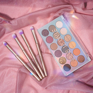 Makeup palette with various shades and four brushes on pink fabric.